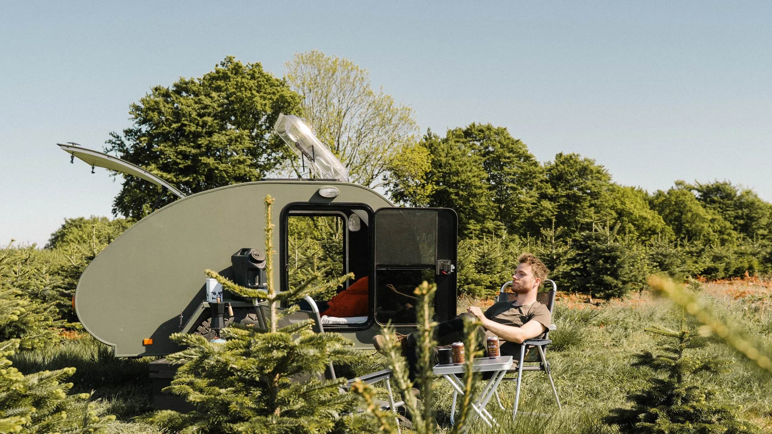 Person sidder i en stol ved en åben campingvogn mellem træer.