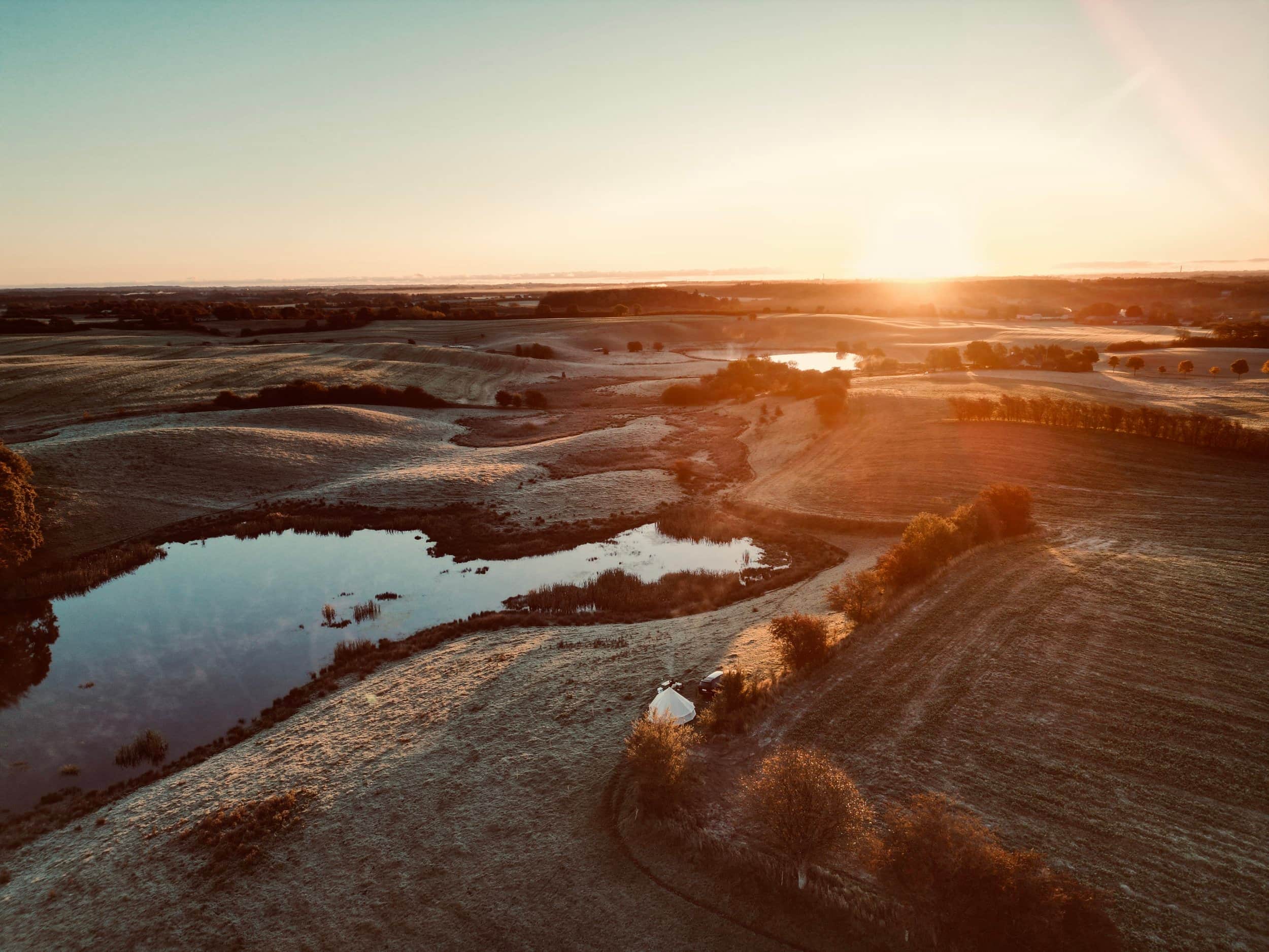Luftfoto af en sø og et telt i landskabet ved solnedgang.