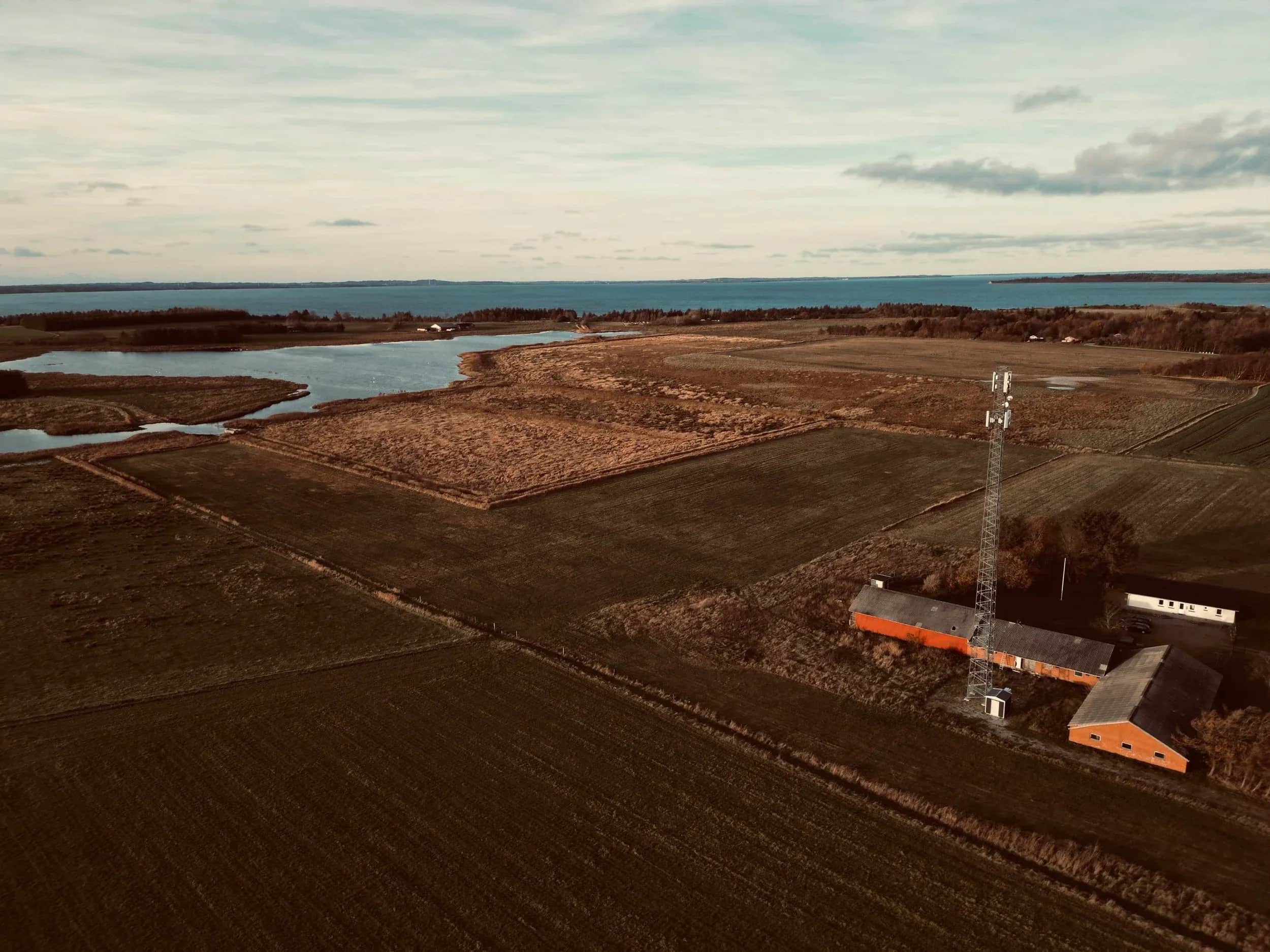 Luftfoto af marker, en fjord og røde bygninger ved en mast.
