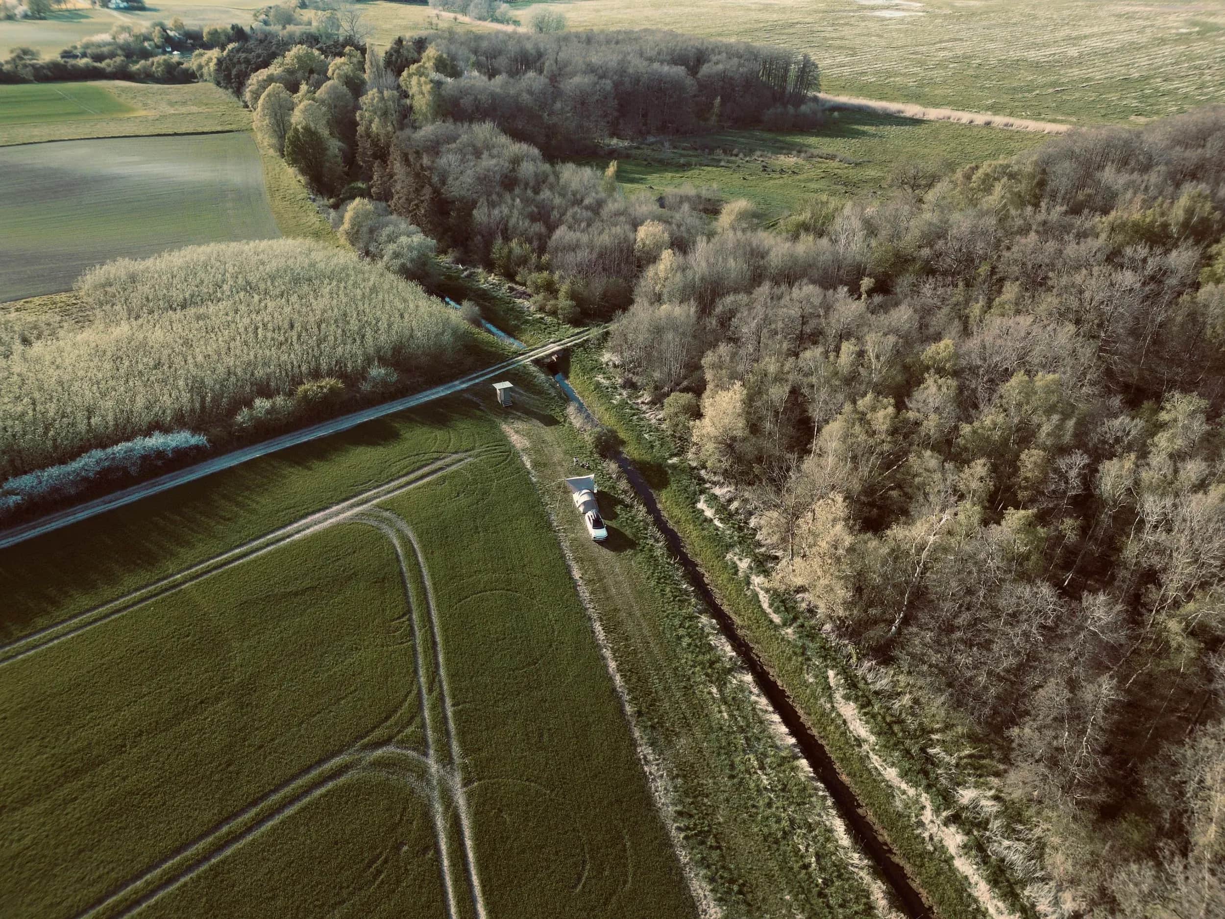 Luftfoto af en bil ved et spor mellem mark og skov.