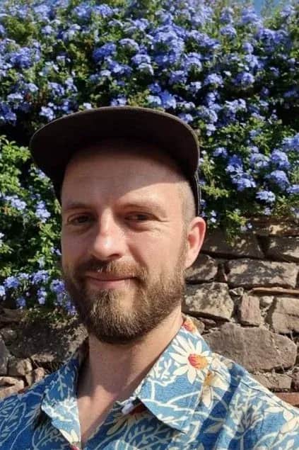 Portræt af en mand med kasket foran blå blomster og en stenmur.