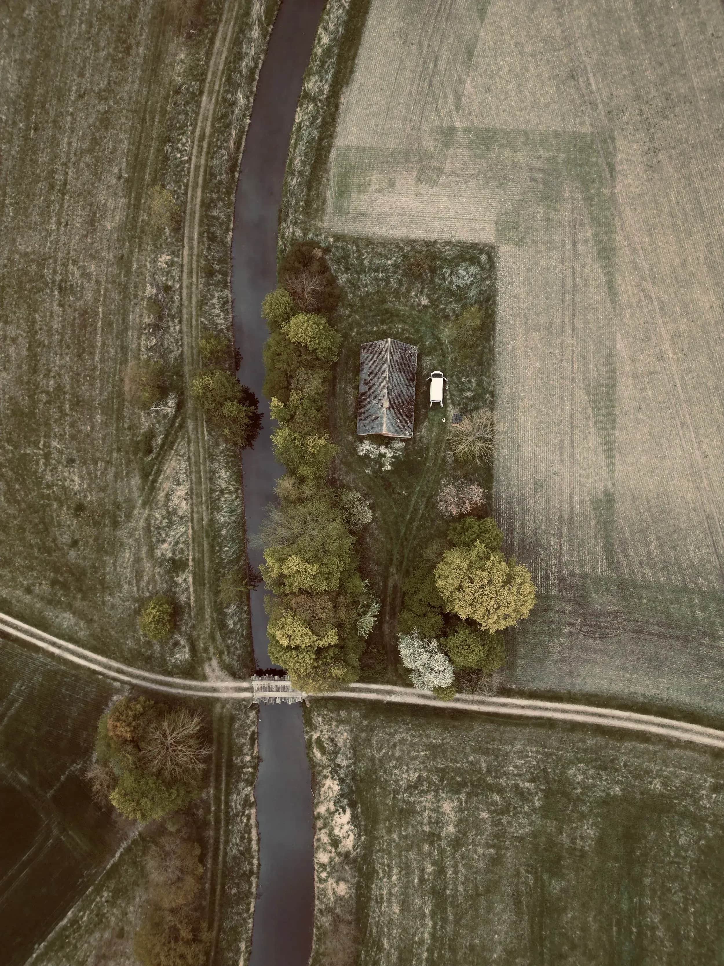 Luftfoto af en å, en vej og et lille hus mellem marker.