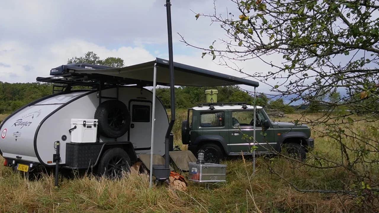 Campingvogn med fortelt og en grøn bil mellem træer.